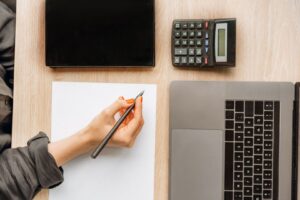 Hand mit Stift neben Laptop, Tablet und Taschenrechner auf einem Schreibtisch